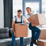 With boxes in hands. Two young movers in blue uniform working indoors in the room.
