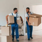 Front view, holding stuff. Two moving service employees in a room.